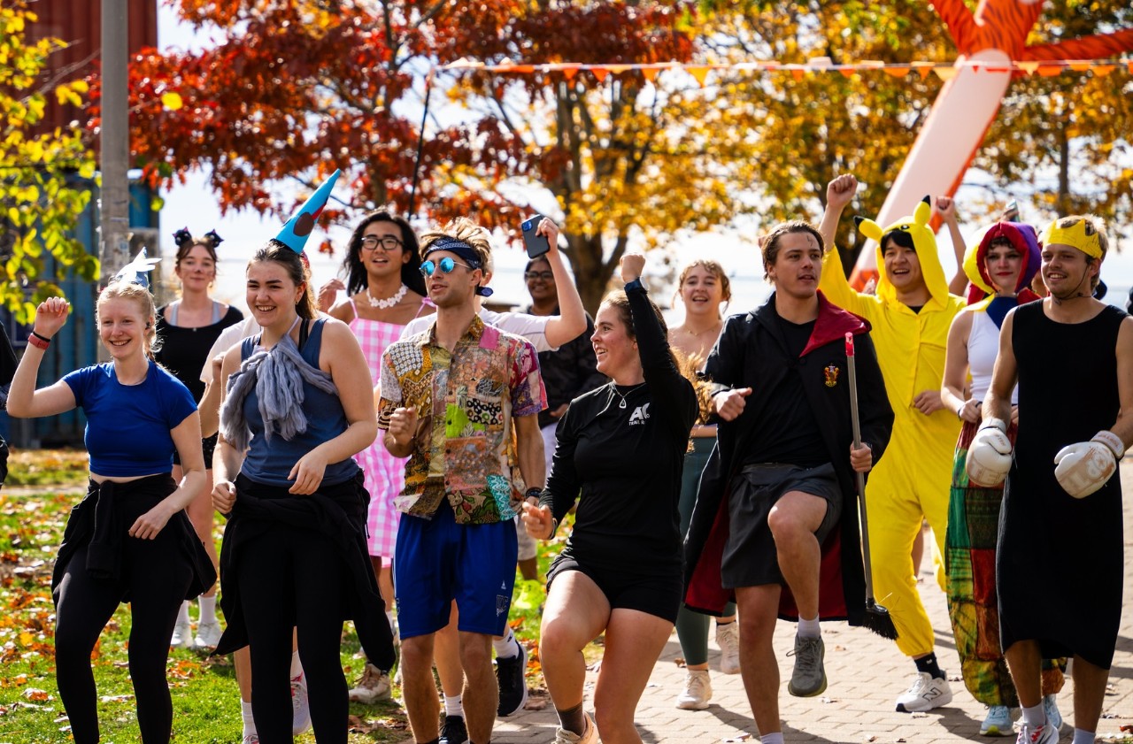 A group of students wearing various costumes and running gear stand outside surrounded by trees with colourful leaves.