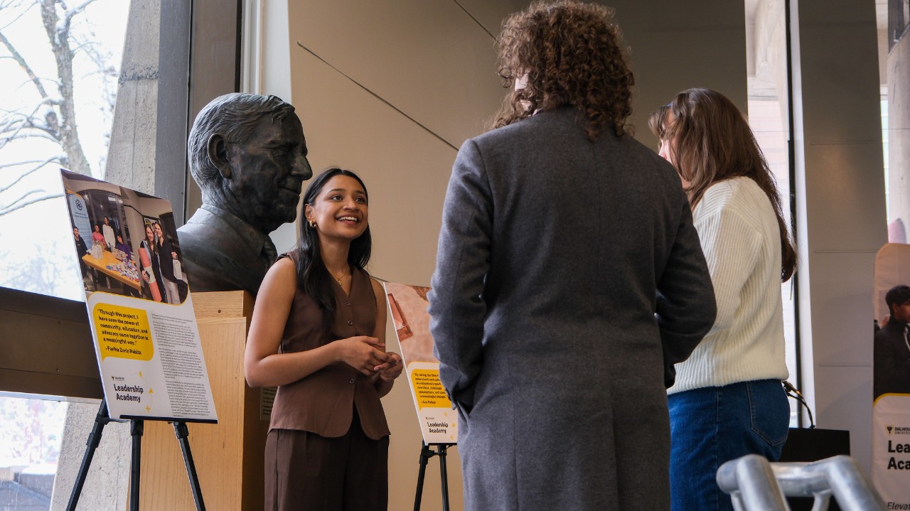 A student wearing a brown vest talks to two other people while standing in front of posters on easels and a large sculpture of a man's head.