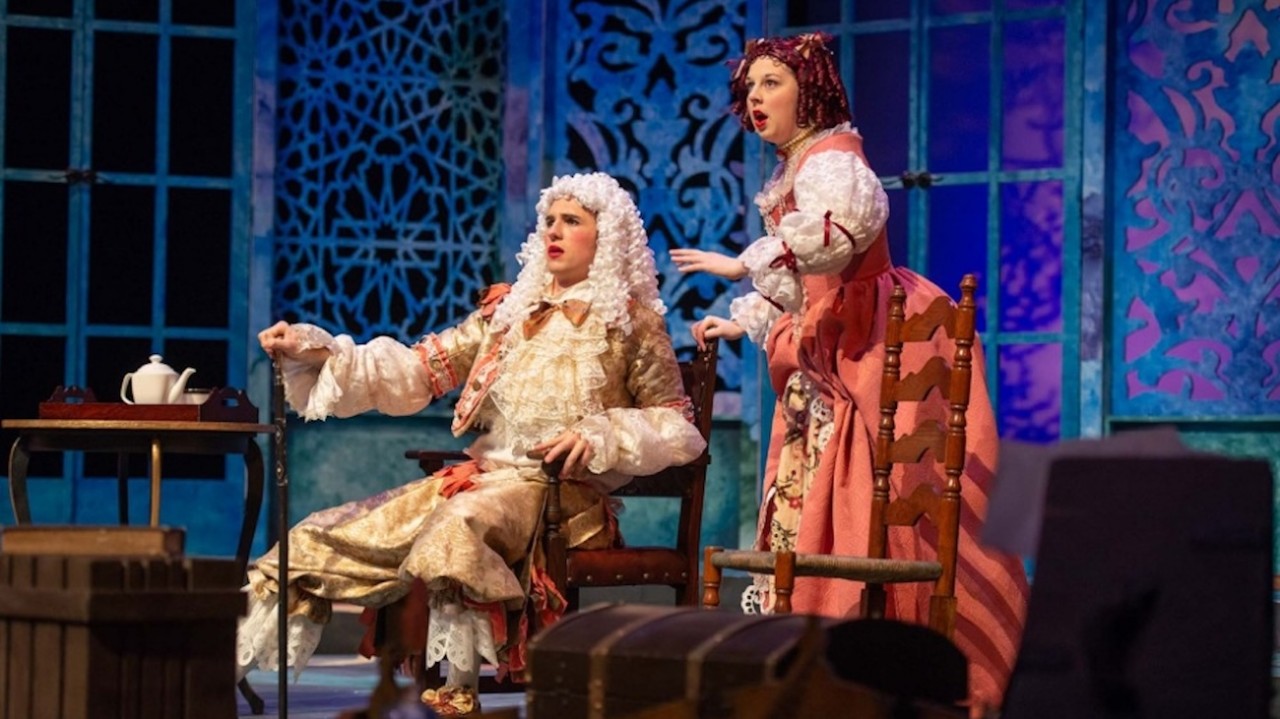 Two actors are on stage, one sitting and the other standing. They are wearing wigs and clothes of the 17th or 18th century.