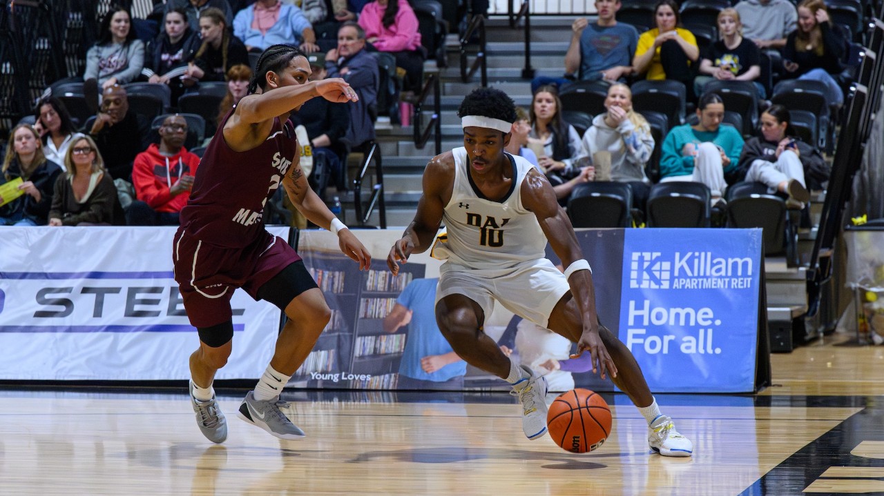A Dalhousie basketball player dribbles the ball on the court as he moves past a defender from Saint Mary's. There are fans in the stands in the background.