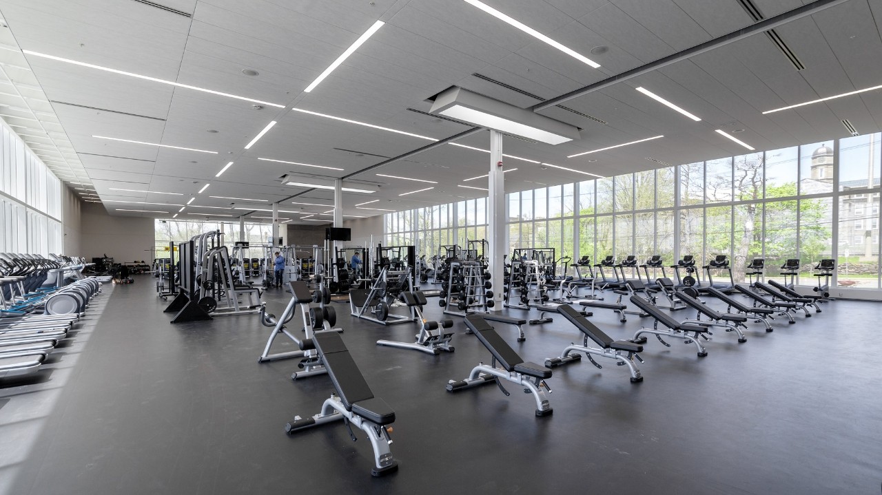 A large open room with a grey floor, white ceiling, and floor to ceiling windows on three sides is filled with various exercise equipment. 