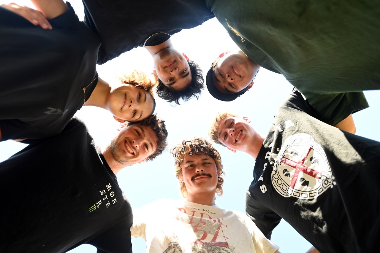 Six students stand in a circle with their heads close together and look down at the camera below.