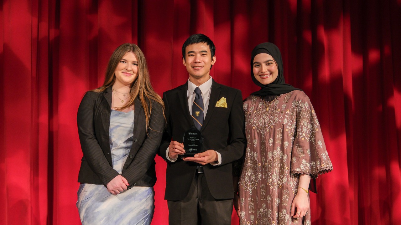 A student wearing a jacket and tie holds a small award in front of him. He is flanked by two women: one in a silver dress and grey jacket; the other wearing a brown dress and black headscarf. They are standing in front of a red curtain with large stars illuminated on it.