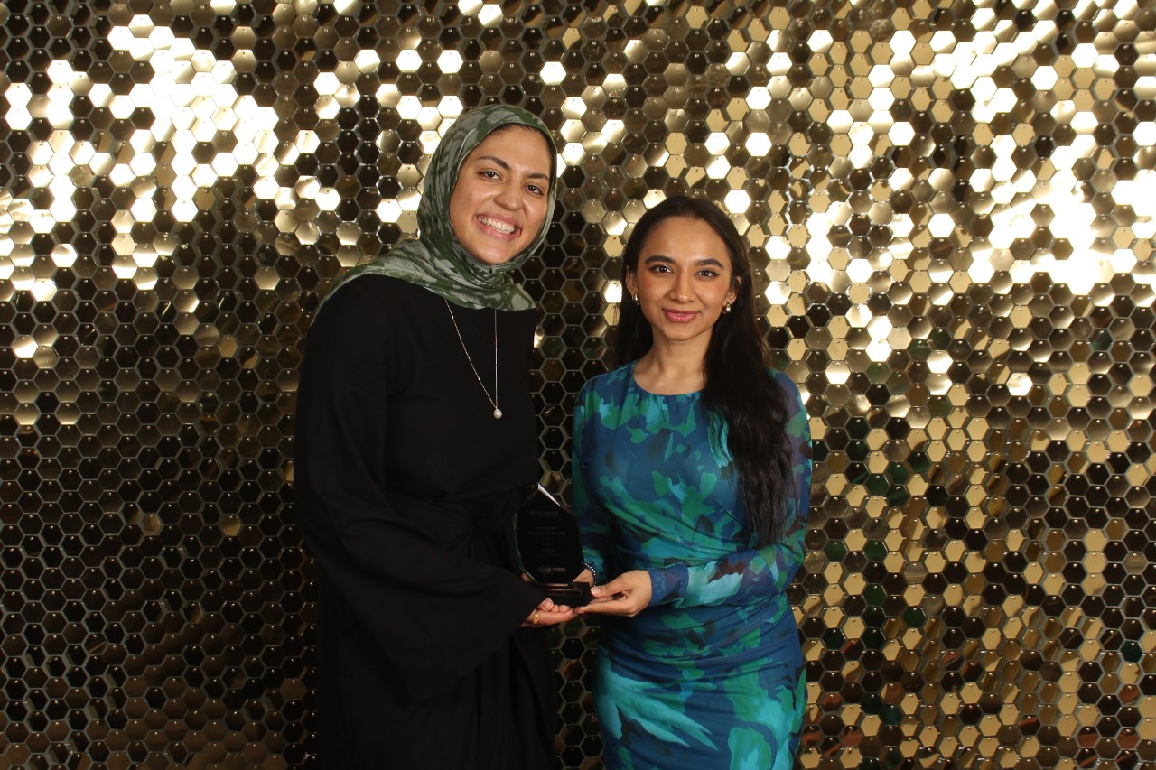 Two students stand in front of a sparkly gold background. One is wearing a black dress and green headscarf. The other is wearing a green and blue dress.