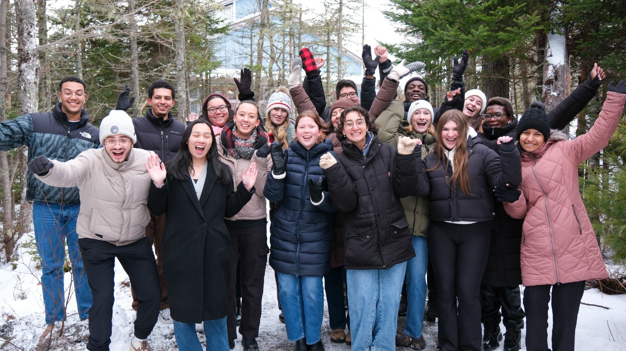 A group of students poses with their arms outstretched in the air while standing in a woodland clearing with snow on the ground.