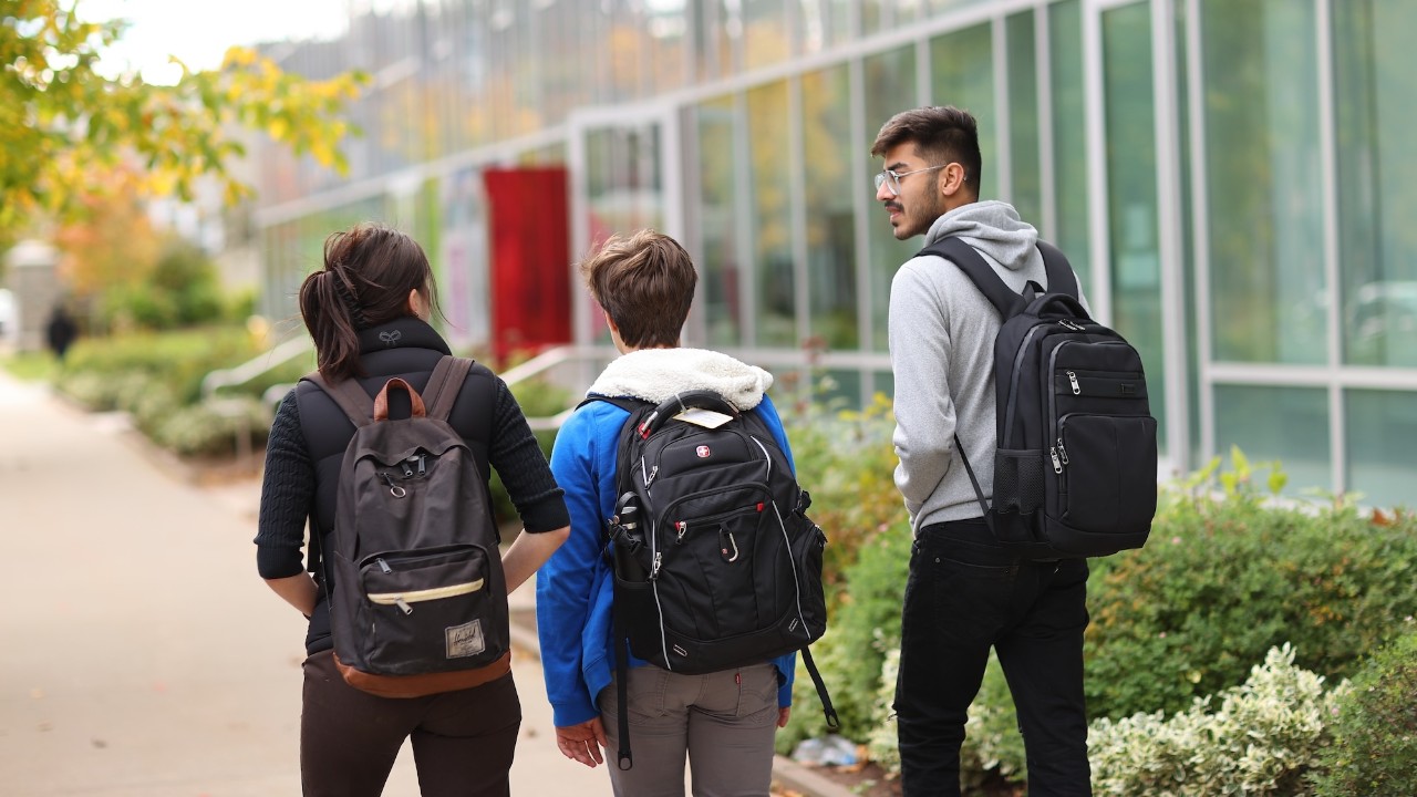 Three students wearing backpacks walk along a sidewalk beside bushes and the glass facade of a building.