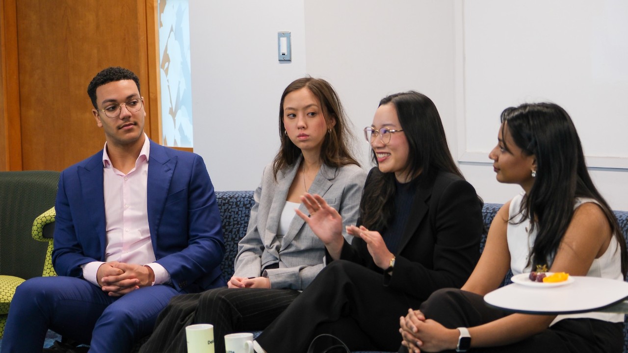 Four students sit beside each other on a large sofa, wearing business clothing. One is talking while the others listen and look at her.