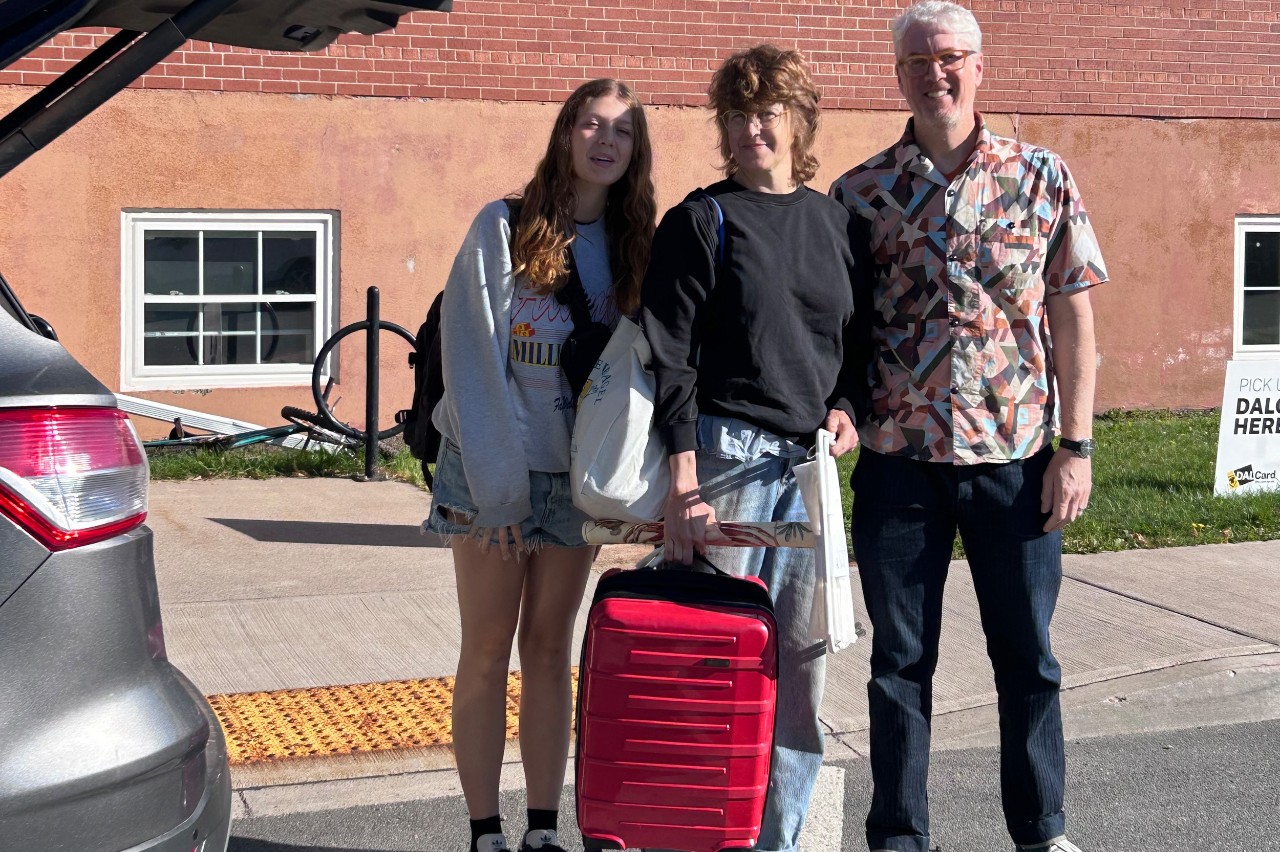A student stands near a vehicle with her parents. Her mother is holding the handle of a red suitcase as she moves into residence. 