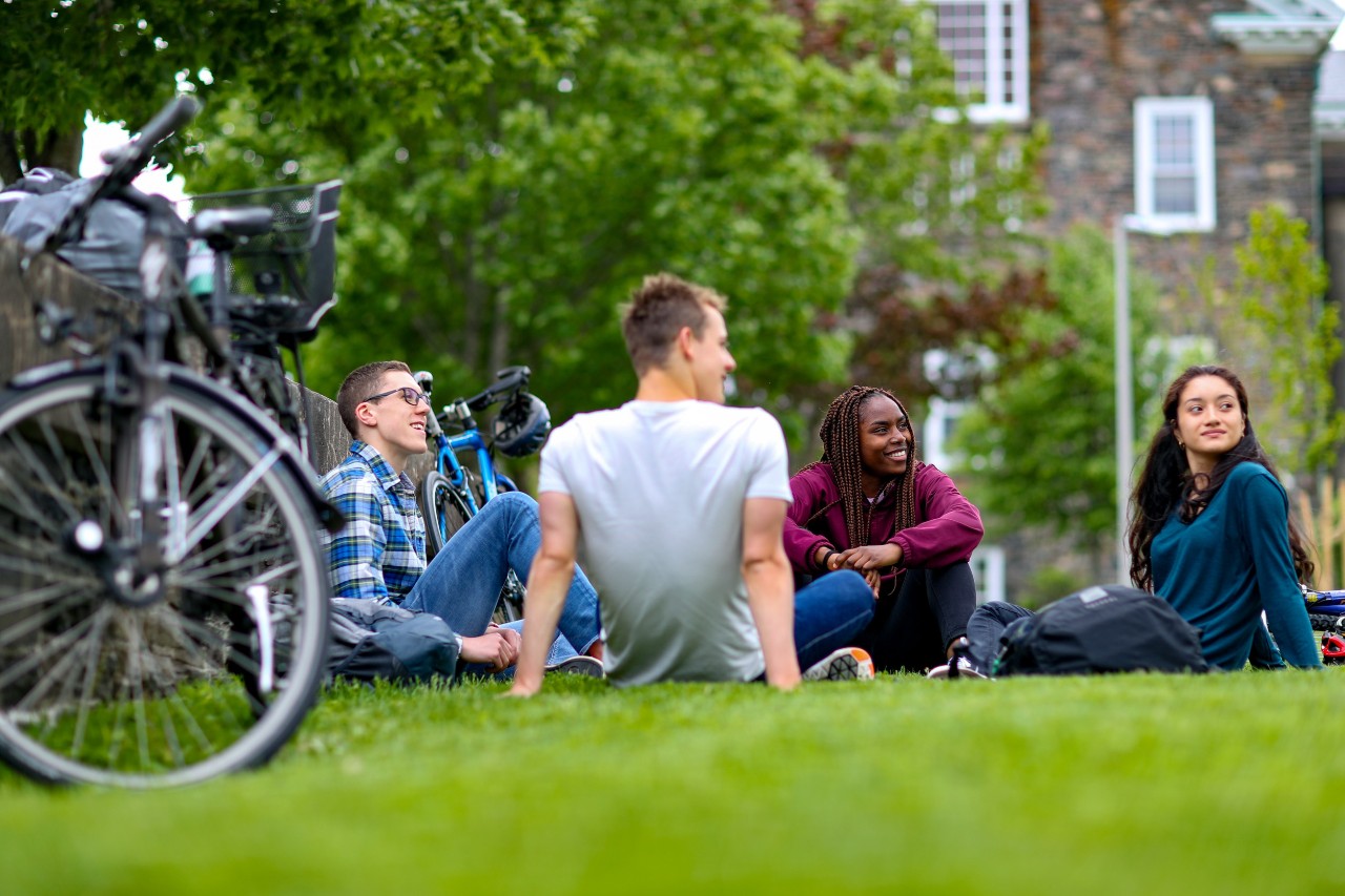 Four students sit on the grass on the Quad at Dalhousie’s Halifax campus on a summer day. Next to them are some bicycles leaning against a concrete wall.  