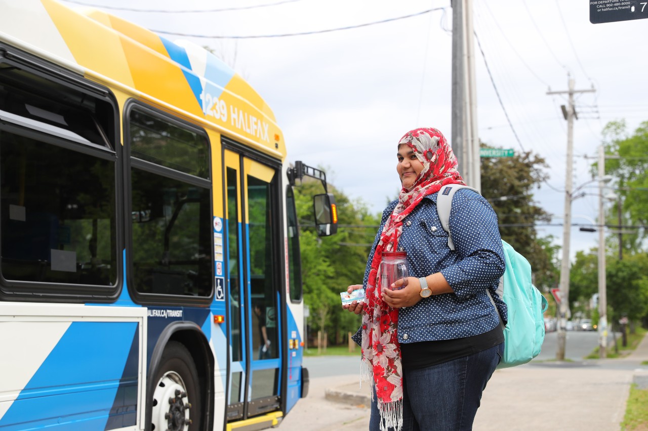 A female student wearing a hijab and a backpack, carrying a glass tumbler, holds her DalCard as a Halifax Transit bus pulls up to the stop. 