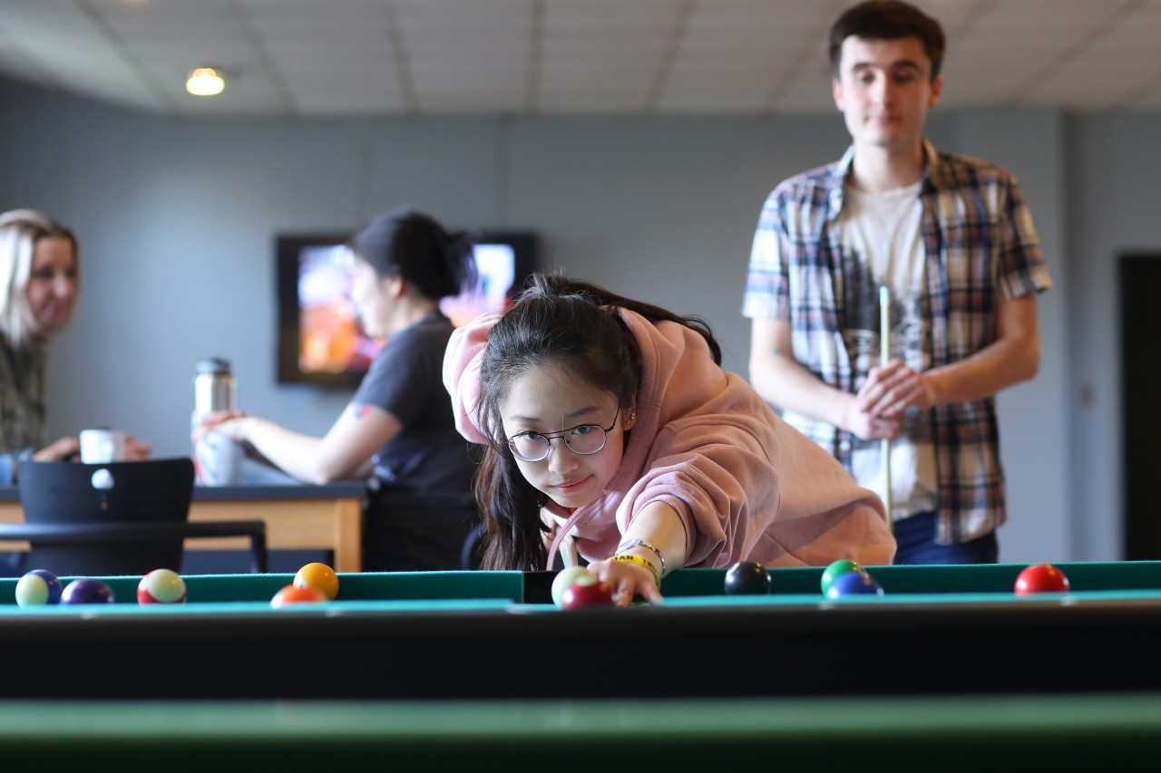 Two students play a game of pool in a residence lounge, while two students are seated at a table behind them talking over coffee.  