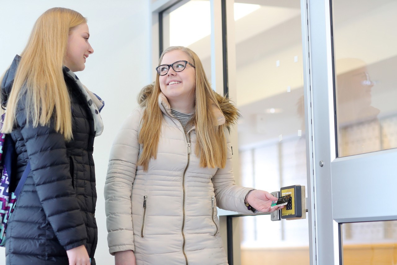 Two female students in winter coats chat as one student taps their DalCard on the entry reader at the front door of Risley Hall.