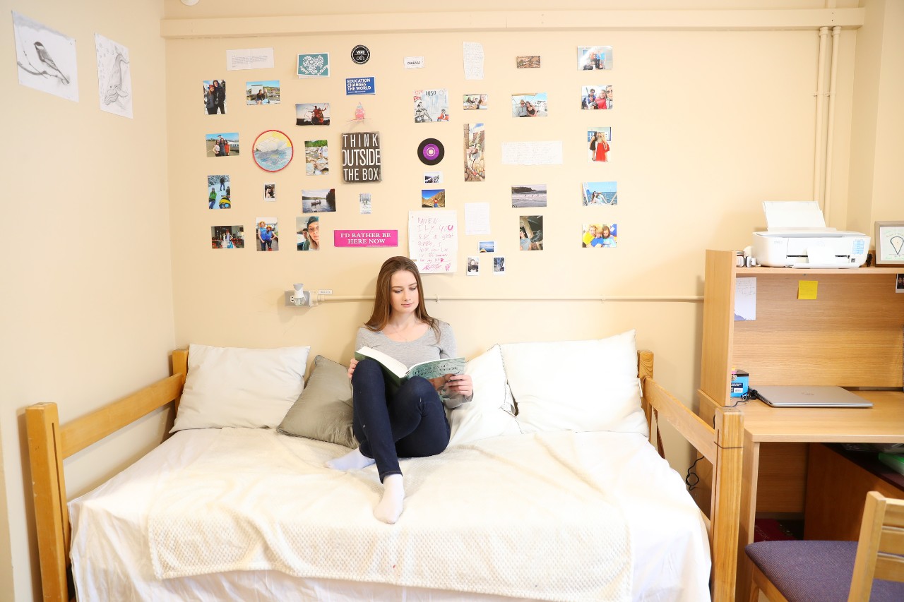A resident sits on her bed in her room in one of our Truro residences studying a textbook. There are photos and posters on the wall and study supplies on her desk. 