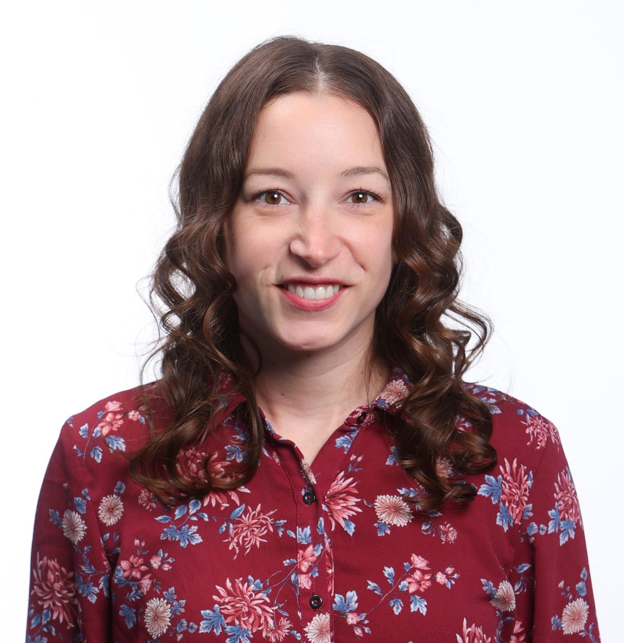 A smiling person with long curly hair and a red floral printed button up shirt
