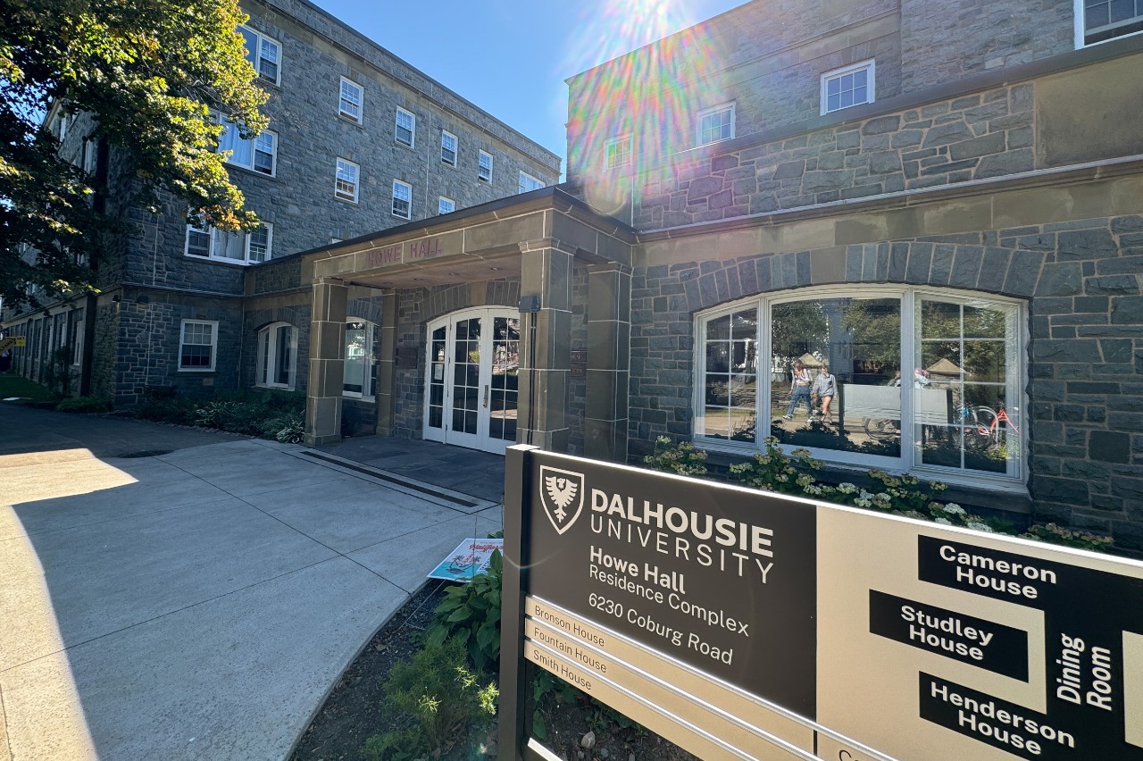 A photo showing the stone-faced front entrance of Howe Hall, including the official Howe Hall sign. Trees are in full bloom and the sky is sunny. 