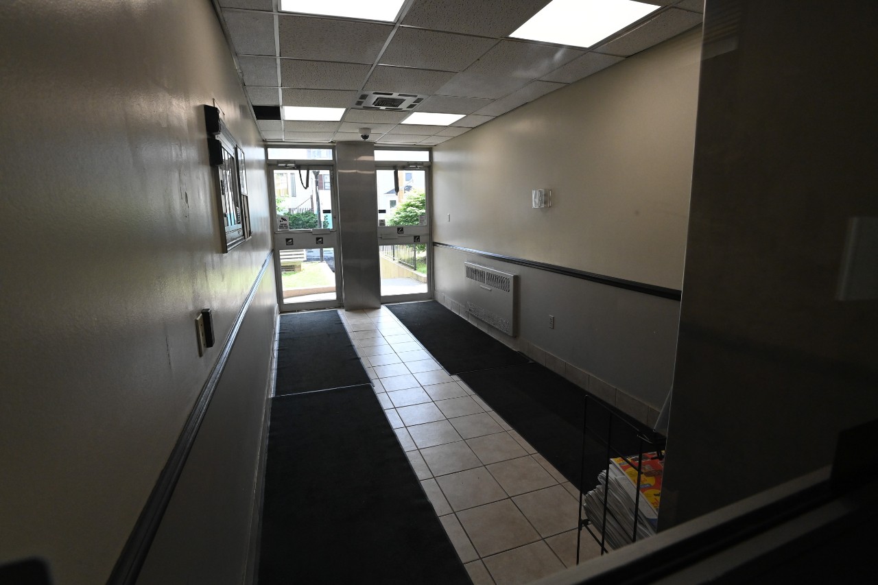 A hallway leads from the front entrance to the main lobby, with a tile floor covered in black carpet runner and 2 glass doors