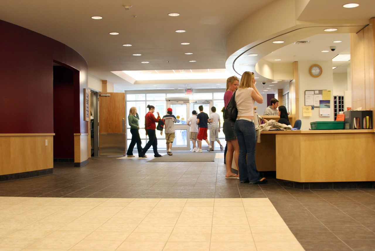 The Risley Hall lobby, bustling with students, with the washrooms and dining hall on the left and front desk on the right, looking toward the front door.