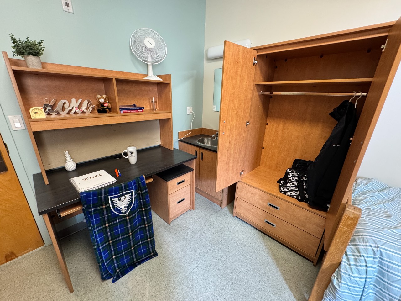 An open wardrobe on one side of a double room in Risley Hall, with some clothing hanging. Next to it is a desk and shared sink.