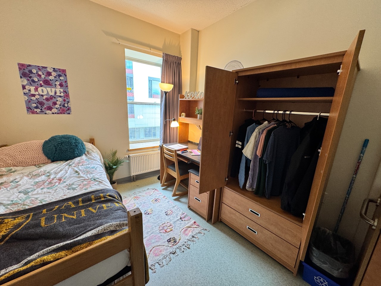 A single room in Risley Hall with an adjustable bed, desk, wardrobe, and a mini fridge. The wardrobe is open revealing shirts hanging inside.