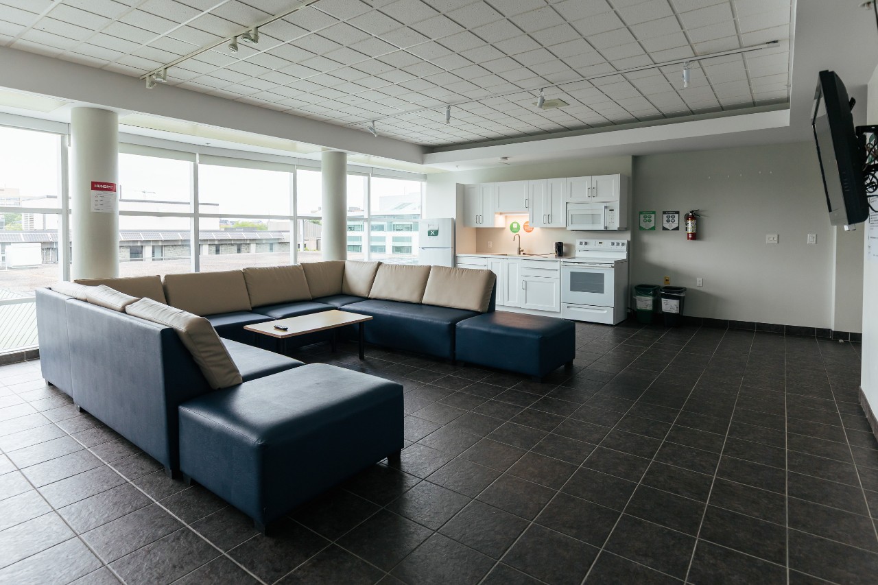 A large lounge on a residence floor in Risley Hall - with a u-shaped sectional sofa, television on the wall, and full kitchen with white cabinets and appliances