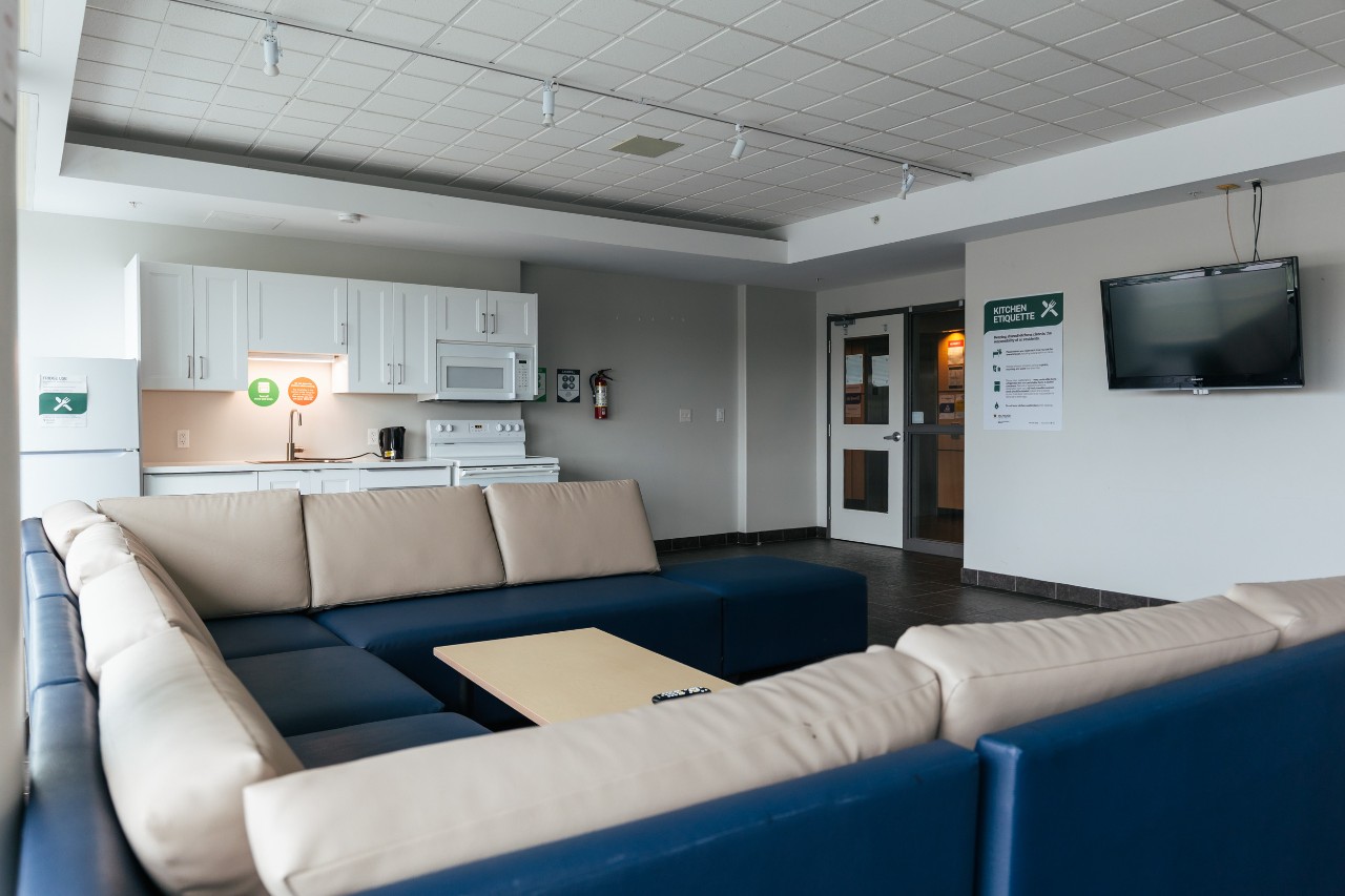 A large lounge on a residence floor in Risley Hall - with a u-shaped sectional sofa, television on the wall, and full kitchen with white cabinets and appliances