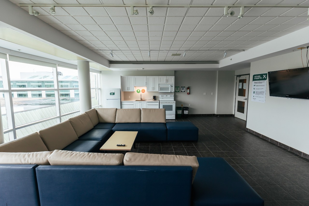 A large lounge on a residence floor in Risley Hall - with a u-shaped sectional sofa, television on the wall, and full kitchen with white cabinets and appliances