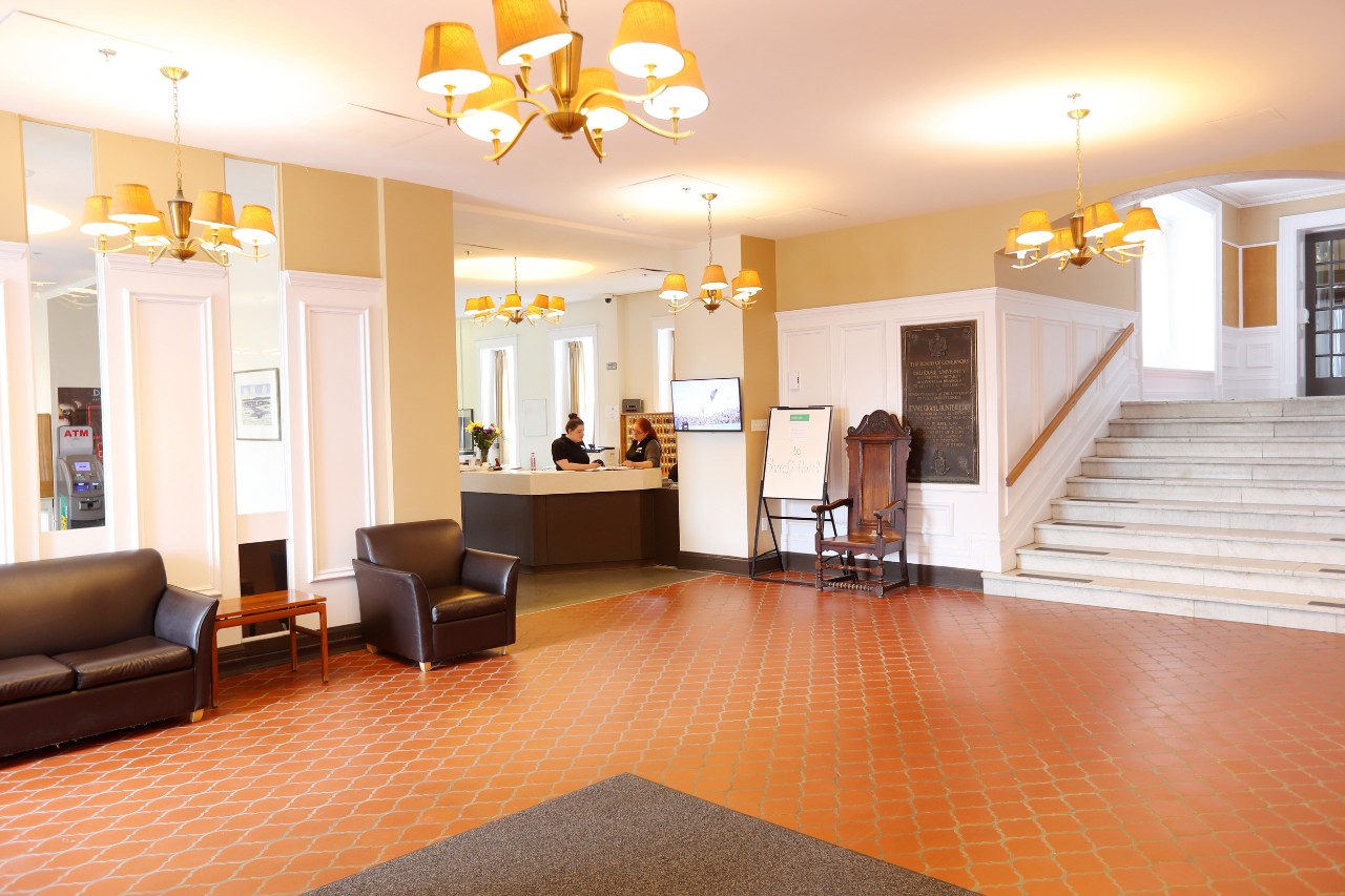 The lobby features comfortable seating, a front desk, and some decorative items. The marble stairs lead toward the dining hall.