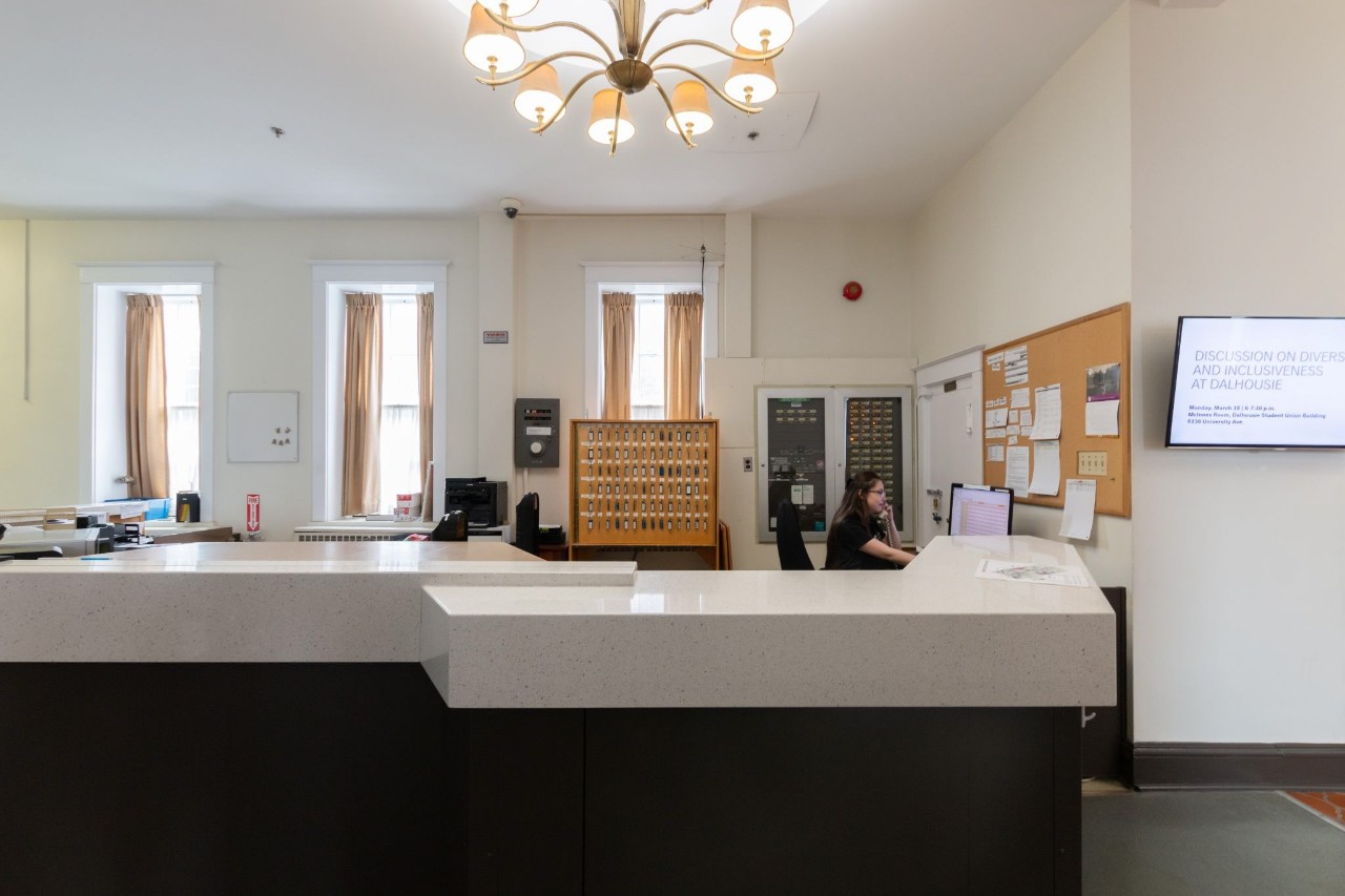 The front desk at Shirreff Hall has white countertops and a dark base. Behind it, a staff member is on the phone and a panel of keys can be seen.