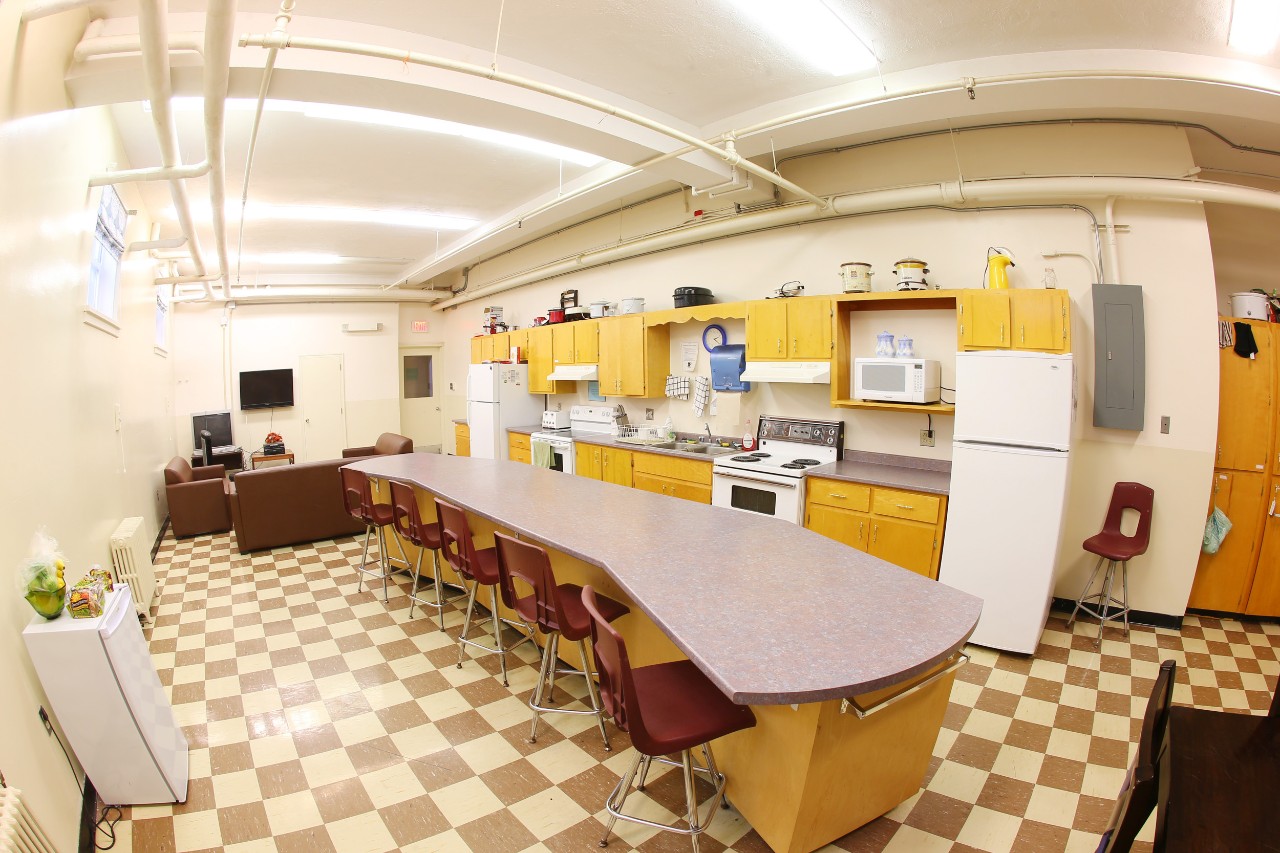 A wide-angle image of the graduate and mature housing full kitchen and lounge – a bright space with light wood cabinets and cream walls and checkered tile floor – with a long island in the middle and comfortable seating area with sofas at the far end. 
