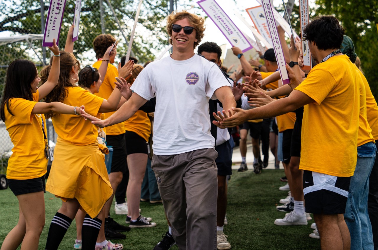 A smiling student wearing a white t-shirt and sunglasses slaps hands with people wearing yellow t-shirts standing in rows on either side of him. They are holding signs in the air with their other hands.