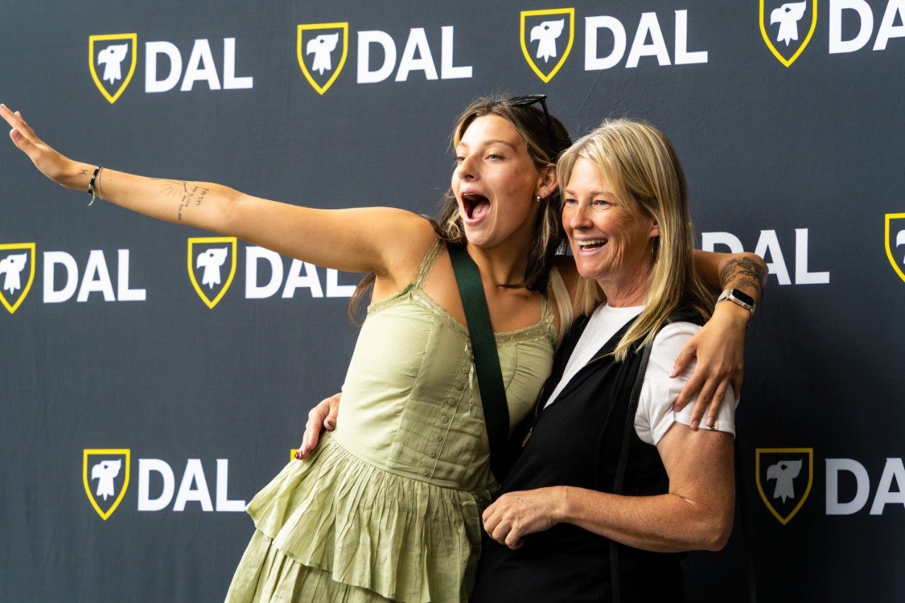 A student and her mother stand with their arms around each other in front of a black backdrop featuring the Dal logo multiple times. They both are smiling and the student has her arm stretched out. 