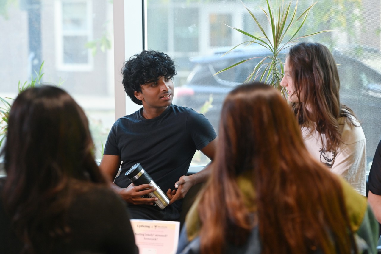 Four students sit together in front of large windows and a green plant. The two in the foreground are facing away to the other two who are talking with each other.