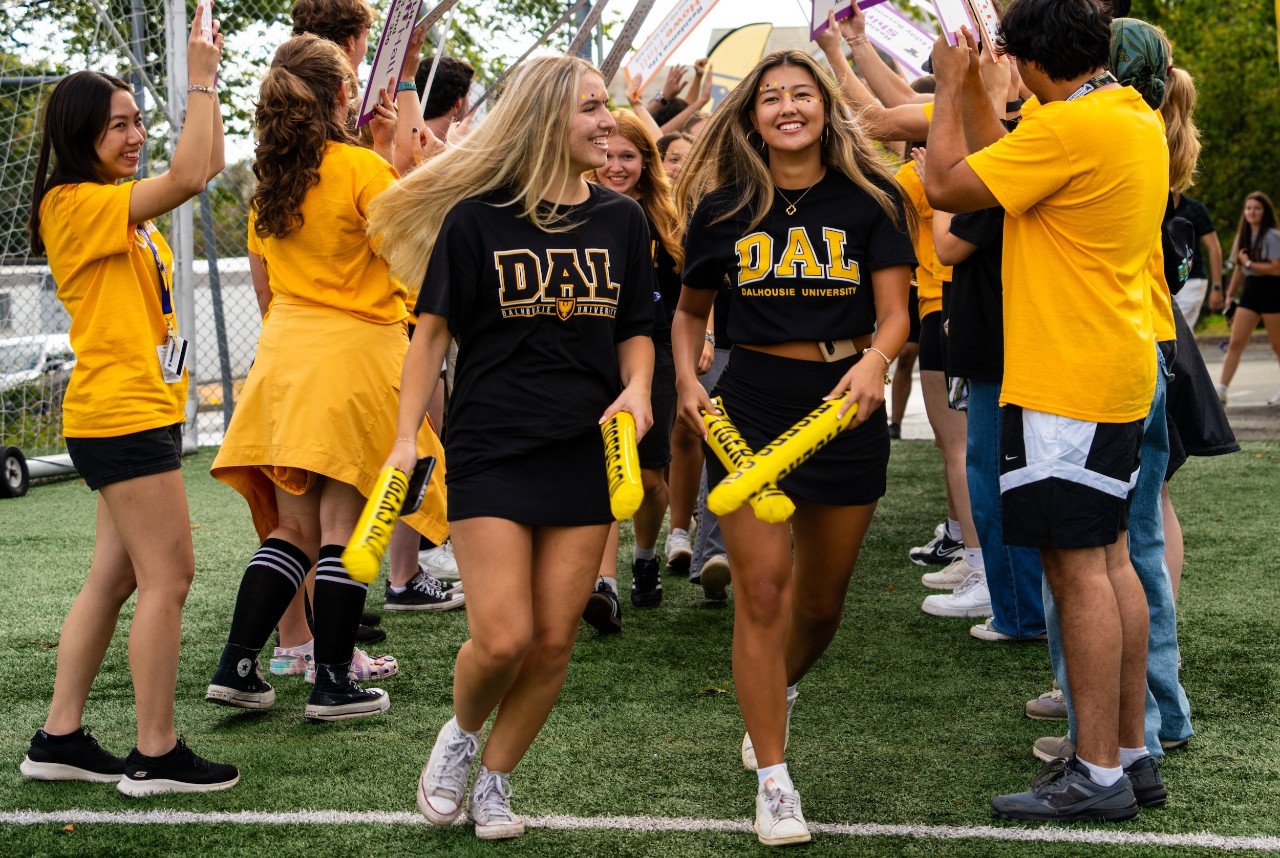 Two female students wearing black t-shirts with Dal written on the front in gold, run through a tunnel of other students wearing gold t-shirts.