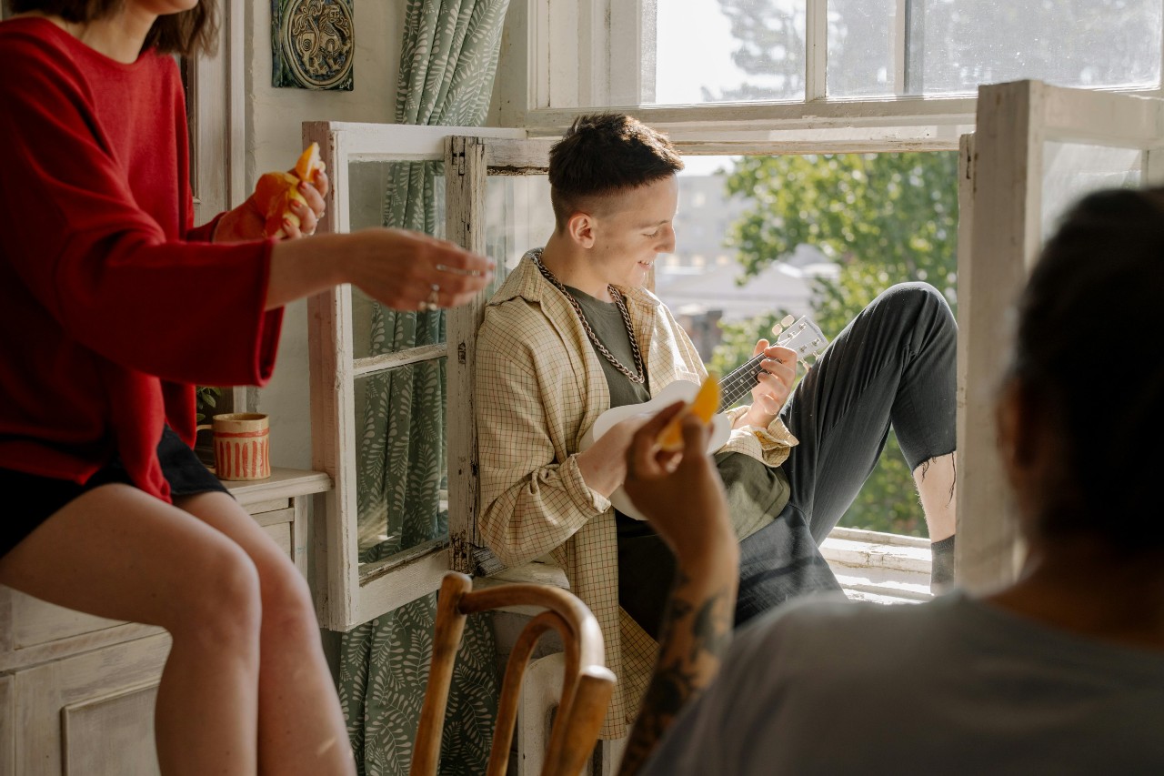 Students laugh and play music near the window of their flat on a sunny day