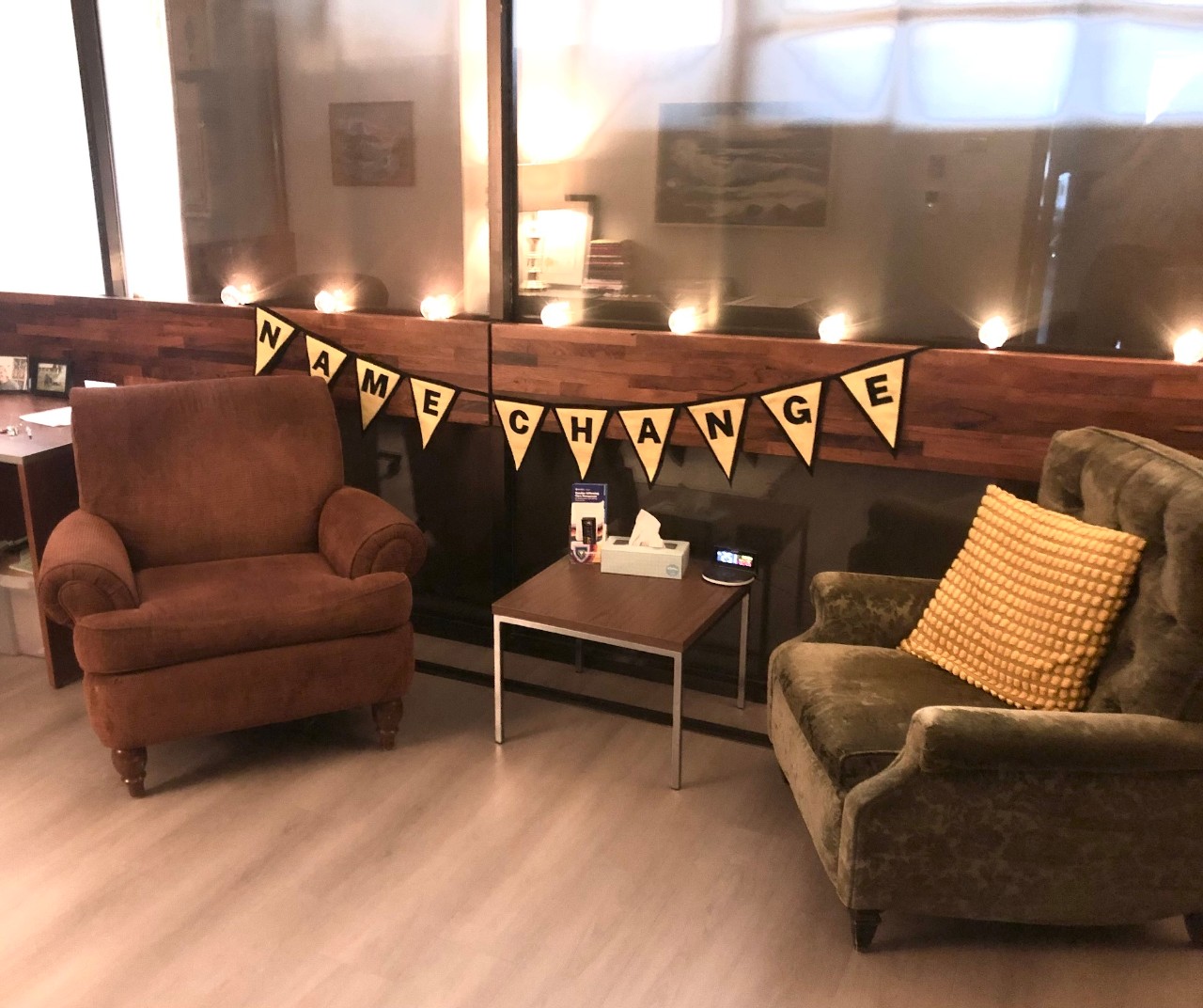 Two comfortable chairs face each other with a small table between them, under a string of yellow and black pennants that say name change.