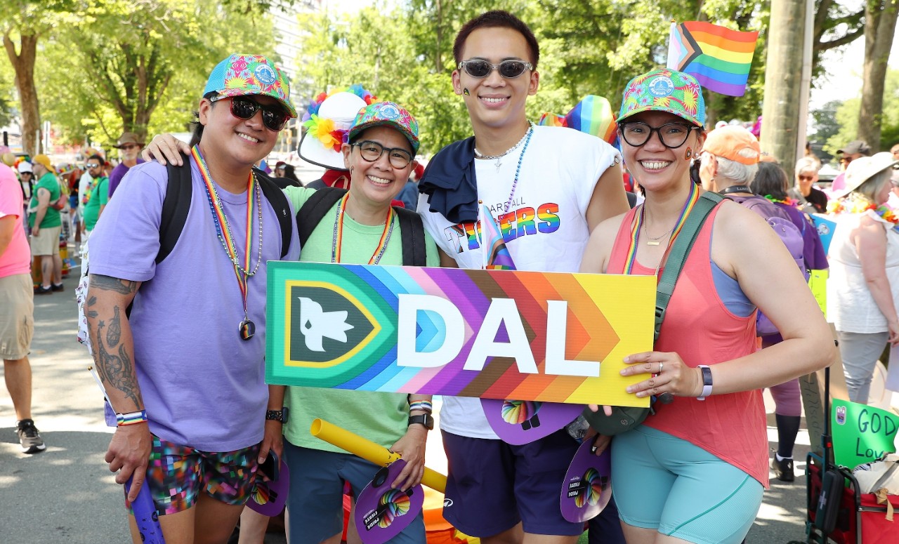 Four young people wearing colourful clothing stand together holding a rainbow-coloured sign that says Dal in front of them.