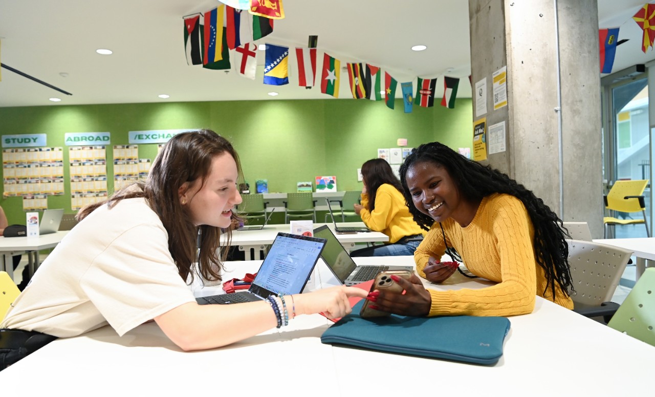 Two students sit across a table from each other, one in a white t-shirt, the other in a yellow sweater. The first one is pointing to the other's mobile phone that she's holding.