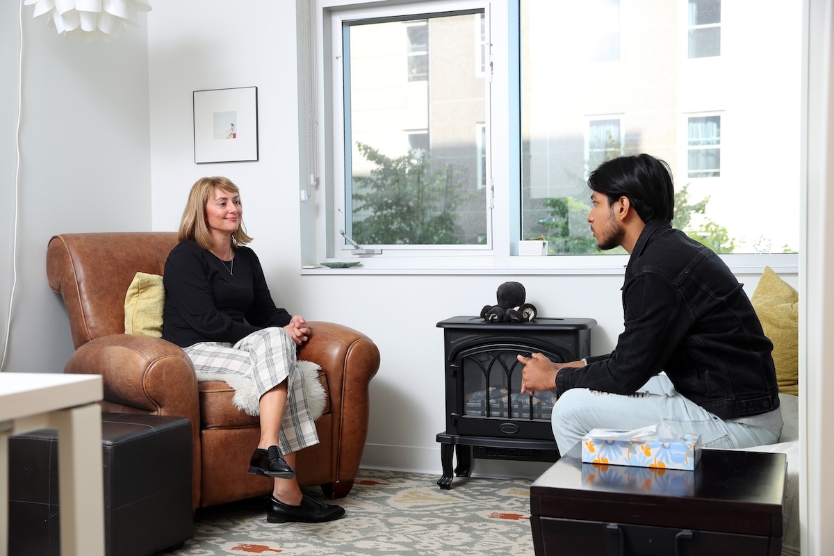 A counsellor sits in a brown chair across from a student sitting in another comfortable chair. There is an electric fireplace beside them.