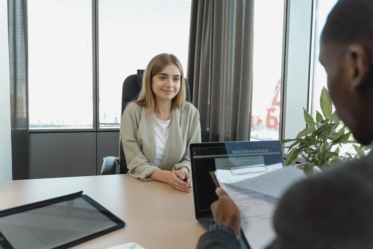 A young woman in business clothes sits across a desk from someone who is looking at her resume. Photo by Tima Miroshnichenko.