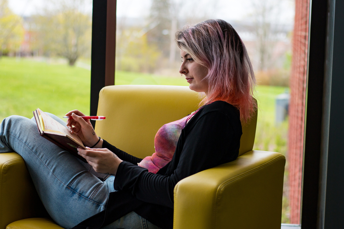 A student with pink dyed hair, wearing a tie-dyed shirt and cardigan, sits in a chair with her legs over the armrest while writing in a notebook.