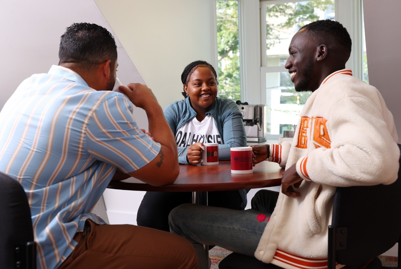 Two students with red coffee mugs sit at a round table and smile with an advisor at the Black Student Advising Centre.