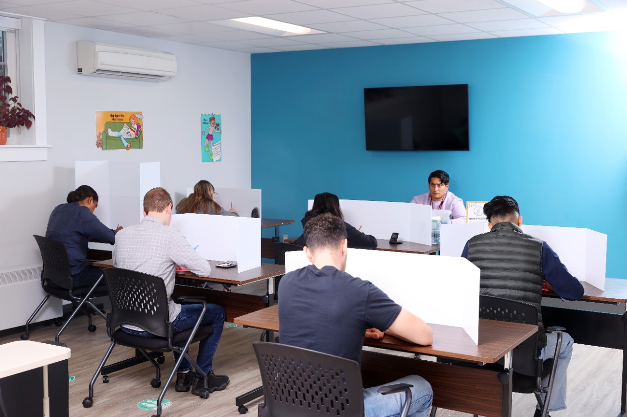 Six students sit and write at separate desks with privacy partitions on them. An invigilator sits in front of a blue wall facing them. 
