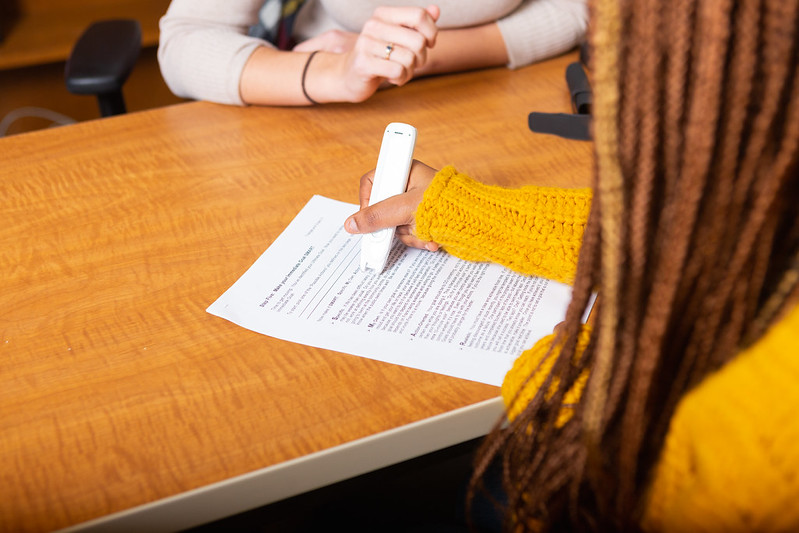 A student with long braids and a yellow top uses an assistive technology tool on a piece of paper. An advisor sits on the other side of the table.