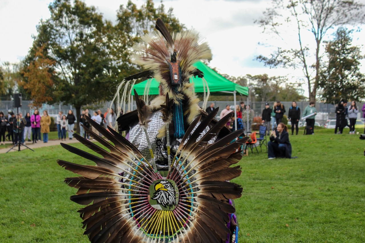 A performer outside at a Ma'wiomi stands facing away with bird feathers decorating their back and head.