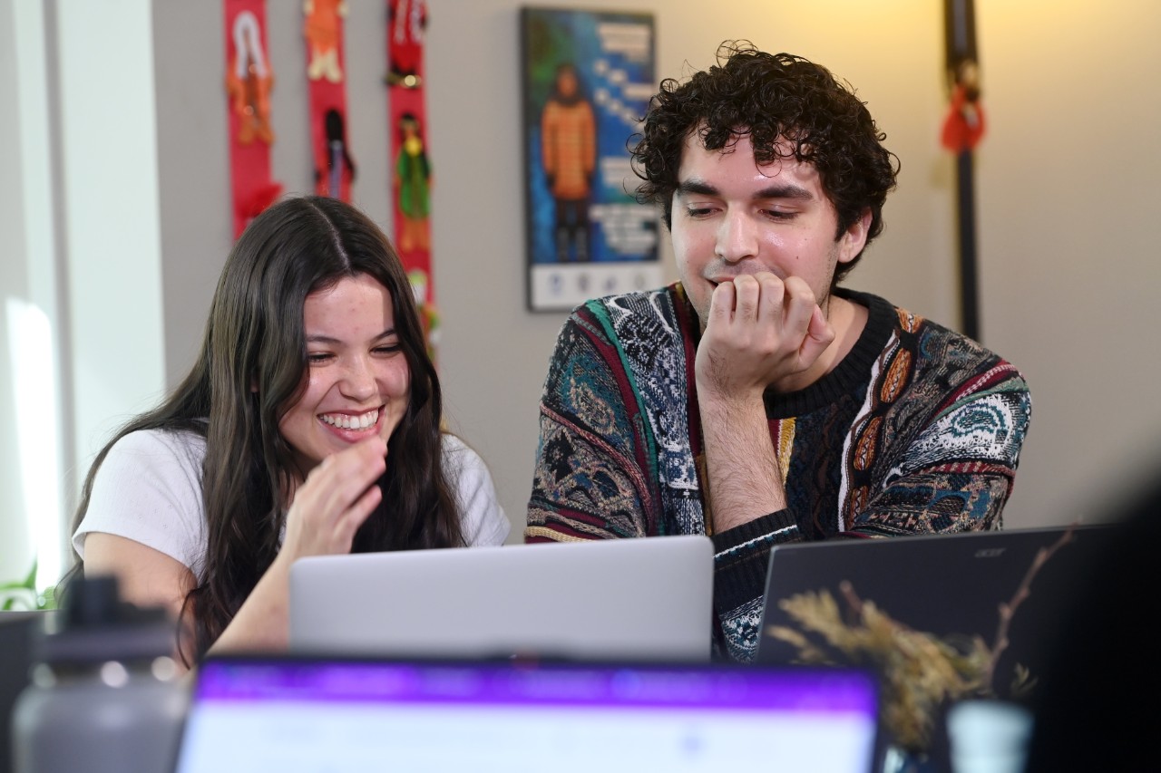 Two students sit and laugh as they look at a laptop screen. In the background is Indigenous artwork on the wall.