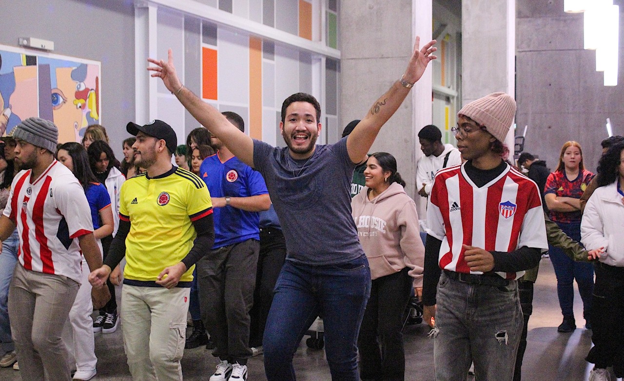 A group of students dances in an open space. Two are wearing red and white striped soccer jerseys, one is in a bright yellow soccer jersey, and one in the middle in a grey shirt has his arms lifted in the air.
