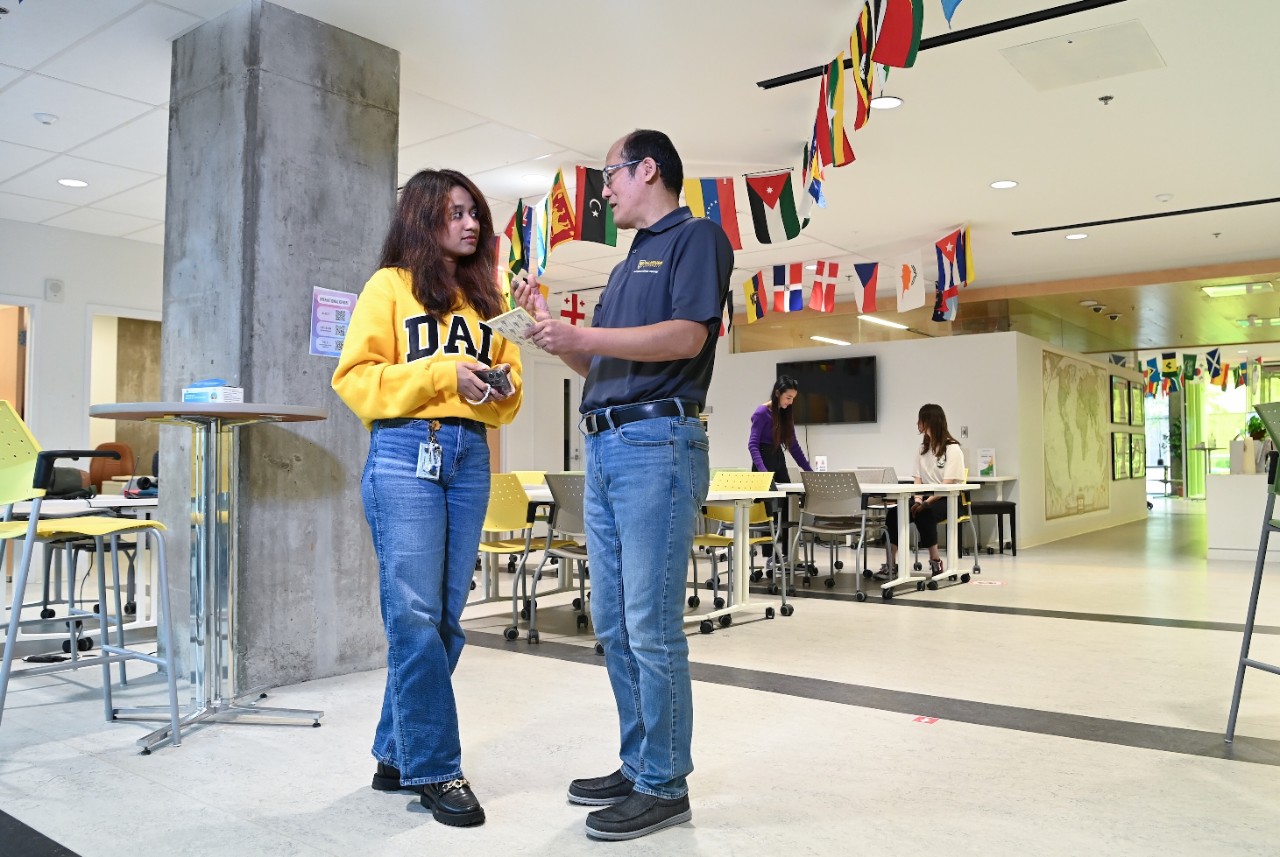 An advisor in jeans and a golf shirt stands and talks with a student wearing a yellow sweater with Dal on the front. They are in a bright room with flags of many countries hanging from the ceiling.