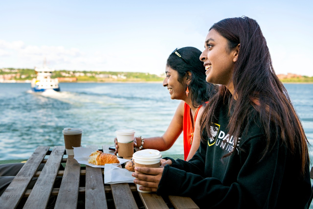Students sitting at a picnic table with to go coffee cups and croissants. A ferry crosses the harbour in the background.