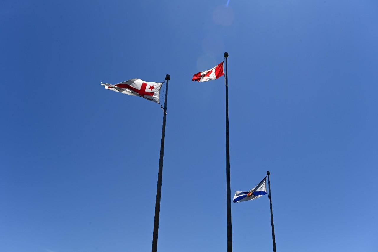 Mi'kmaq, Canada, and Nova Scotia flags fly on three flagpoles in front of a bright blue sky.