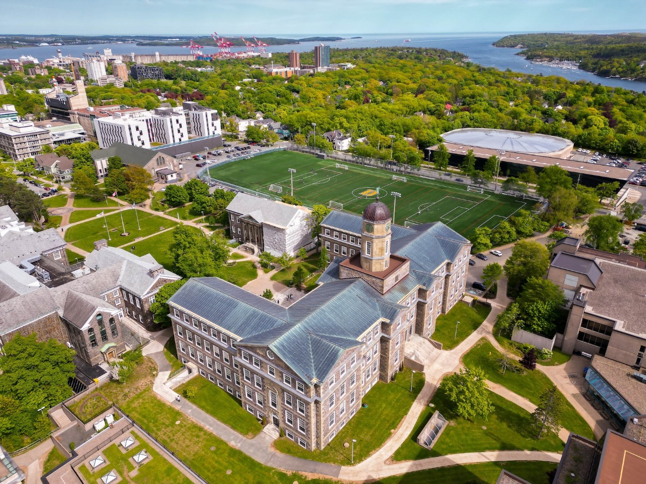 An aerial view of Dalhousie's Studley Campus with an old stone building in the foreground, sports field in the middle, and trees and ocean in the background.